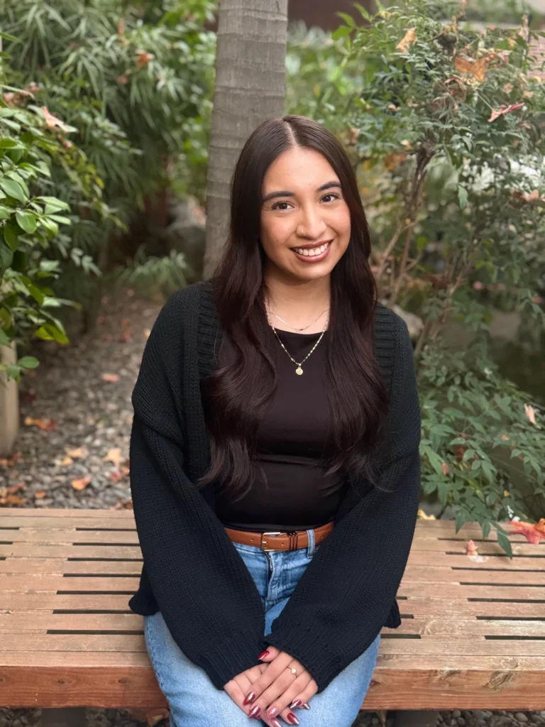 Young woman smiling outdoors in casual attire, surrounded by greenery.